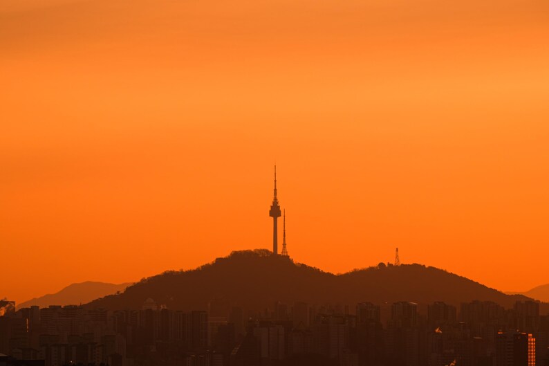 Puede incluir: Una silueta de la torre N Seoul sobre la cima de una monta&ntilde;a contra un cielo de atardecer naranja vibrante y s&oacute;lido. Los contornos oscuros de la torre y las colinas son visibles sobre la tenue silueta de una ciudad en el primer plano.