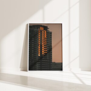 May include: A framed vertical wall art print featuring a high-rise building at sunset. The skyscraper is dark gray with a vertical column of windows reflecting bright orange sunlight against a muted, warm-toned sky during the golden hour.