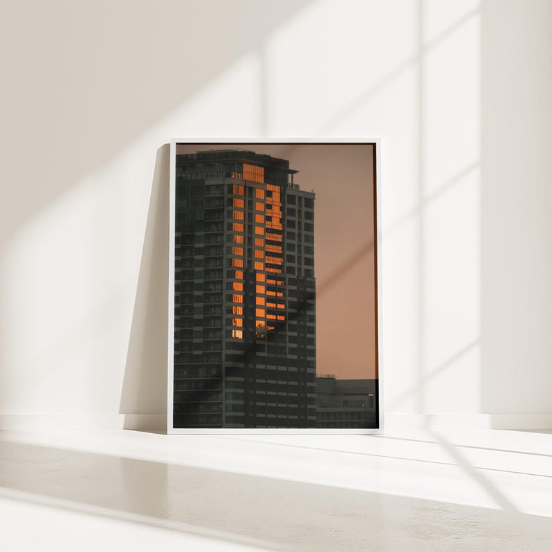 May include: A framed vertical wall art print featuring a high-rise building at sunset. The skyscraper is dark gray with a vertical column of windows reflecting bright orange sunlight against a muted, warm-toned sky during the golden hour.
