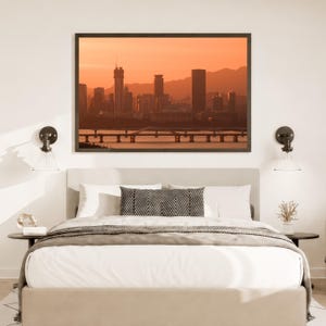 May include: A bedroom featuring a bed with white bedding and a black and white chevron patterned throw pillow, centred beneath a framed landscape print of a city skyline at sunset with a bridge over water, flanked by two wall-mounted lamps.