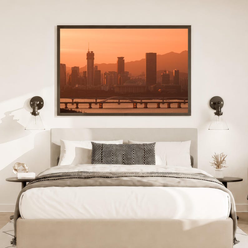 May include: A bedroom featuring a bed with white bedding and a black and white chevron patterned throw pillow, centred beneath a framed landscape print of a city skyline at sunset with a bridge over water, flanked by two wall-mounted lamps.