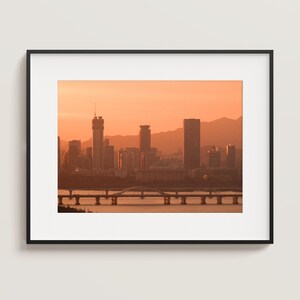 May include: A framed wall art print featuring a city skyline silhouette during a sunset with orange hues. The scene displays multiple skyscrapers, a bridge spanning across a river, and distant mountains under a warm, glowing sky.