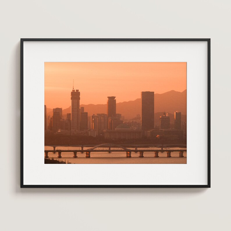 May include: A framed wall art print featuring a city skyline silhouette during a sunset with orange hues. The scene displays multiple skyscrapers, a bridge spanning across a river, and distant mountains under a warm, glowing sky.