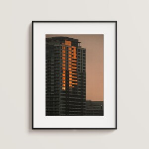 May include: A framed wall art print featuring a high-rise building at sunset. The skyscraper has a dark gray facade with a vertical section of windows reflecting bright orange sunlight against a muted, soft-toned evening sky background.