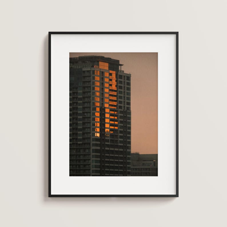 May include: A framed wall art print featuring a high-rise building at sunset. The skyscraper has a dark gray facade with a vertical section of windows reflecting bright orange sunlight against a muted, soft-toned evening sky background.