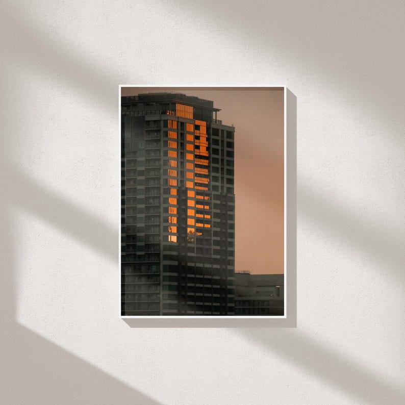 May include: A tall skyscraper captured at sunset, featuring a dark gray facade with a vertical section of windows reflecting bright orange light against a muted, soft-toned sky, creating a high-contrast architectural scene in a vertical frame.