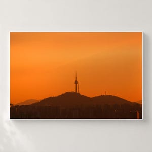 Puede incluir: Una impresi&oacute;n de arte mural enmarcada que presenta la silueta de la torre N Seoul sobre una cresta monta&ntilde;osa contra un cielo naranja vibrante al atardecer. El primer plano muestra los contornos oscuros de edificios bajo las colinas.