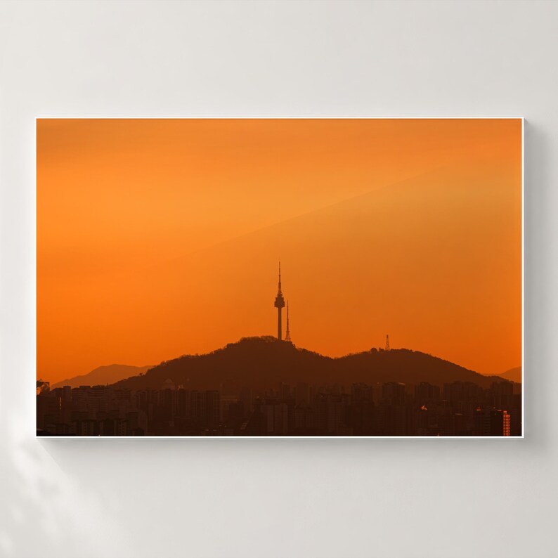 Puede incluir: Una impresi&oacute;n de arte mural enmarcada que presenta la silueta de la torre N Seoul sobre una cresta monta&ntilde;osa contra un cielo naranja vibrante al atardecer. El primer plano muestra los contornos oscuros de edificios bajo las colinas.