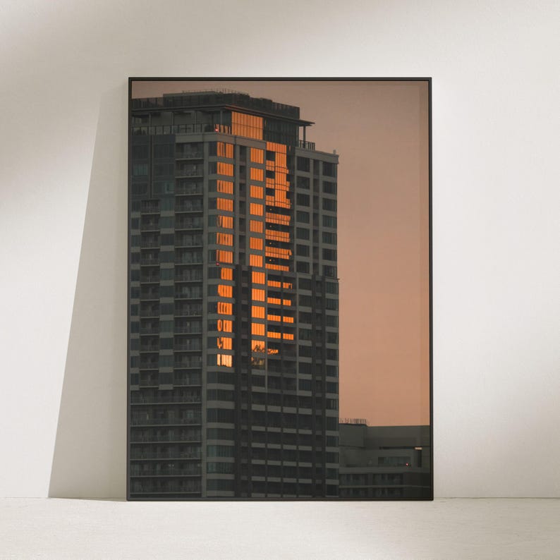 May include: A framed vertical wall art print depicting a tall skyscraper at sunset. The building features a grid of dark gray windows, with a central column of windows reflecting a bright, warm orange light against a muted, dusky sky background.