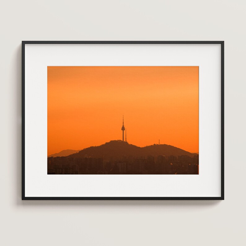Puede incluir: Una fotograf&iacute;a de paisaje enmarcada que muestra la silueta de la torre N Seoul sobre una monta&ntilde;a contra un cielo naranja vibrante al atardecer. La imagen est&aacute; en un marco negro fino con un borde blanco, mostrando la silueta de la ciudad.