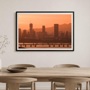 May include: A framed landscape print featuring a city skyline silhouette against a vibrant orange sunset sky. The scene includes tall skyscrapers, a long bridge spanning across the water, and distant mountains, all rendered in warm monochromatic tones.