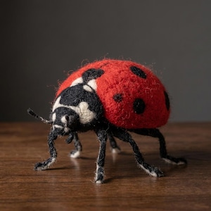 May include: A needle-felted ladybug figurine crafted from wool. The insect features a bright red domed back with distinct black circular spots, a black head with white markings, and six thin black legs extending from the underside.
