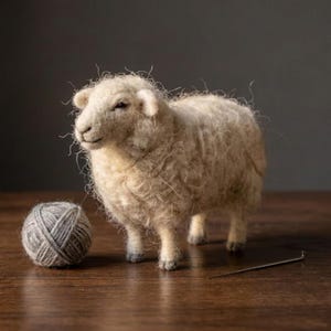 Op de afbeelding: Een naaldgevilte, crèmekleurige schaapfiguur met een getextureerd, wollig uiterlijk, staand naast een kleine bol grijze wol en een enkele metalen naainaald. Het schaap heeft donkere ogen en kleine, gedetailleerde hoeven aan de vier poten.