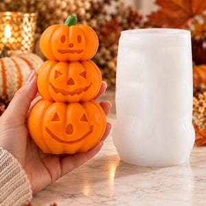 Puede incluir: Una pila de tres calabazas naranjas tipo jack-o'-lantern con caras talladas y un tallo verde, junto a un molde de silicona blanco translúcido utilizado para crear la forma de la pila de calabazas.