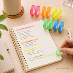 Puede incluir: Un cuaderno de espiral con planes diarios, objetivos y notas escritos a mano, acompañado de diez rollos de cinta washi de colores rosa, naranja, amarillo, verde y azul. Un bolígrafo blanco descansa cerca y una mano aplica una tira de cinta verde.