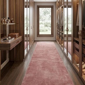 Puede incluir: Un vestidor con armarios de madera oscura con puertas de cristal, una alfombra rosa en el centro sobre un suelo de madera oscura y una zona de tocador con soportes de metal dorado, frascos de perfume y una caja de madera, frente a una ventana.