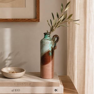 Può includere: Un alto vaso in ceramica a forma di bottiglia con un manico laterale, caratterizzato da un design a blocchi di colore rosa tenue, marrone scuro e uno smalto turchese colante sul collo. Un sottile ramo d'ulivo verde è inserito nell'apertura.