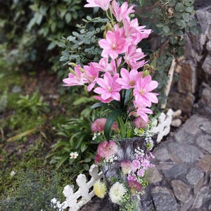 Könnte beinhalten: Ein Blumenarrangement mit hohen rosa Lilien, grünen Stielen und Blättern, akzentuiert durch kleinere rosa, weiße und gelbe kugelförmige Blüten, alles in einem dunklen, strukturierten, rindenartigen zylindrischen Gefäß.
