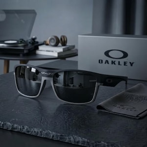May include: Matte black and grey Oakley sunglasses featuring dark reflective lenses and the Oakley logo on the temple. The sunglasses are positioned next to a matching grey microfibre cleaning cloth and a branded Oakley box with the logo printed on it.