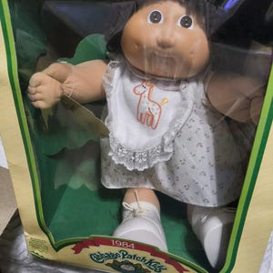 Pode incluir: Uma boneca Cabbage Patch Kids de 1984 com cabelo escuro e rosto redondo, vestindo um vestido branco com um pequeno padrão floral e um babador branco com renda e um gráfico de girafa laranja. A boneca usa sapatos brancos dentro da caixa.