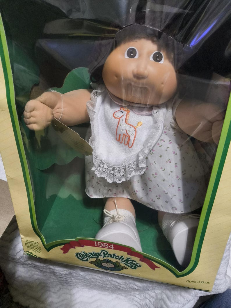 May include: A 1984 Cabbage Patch Kids doll with dark hair and a round face, wearing a white dress with a small floral pattern and a white lace-trimmed bib featuring an orange giraffe graphic. The doll wears white shoes and is inside its original box.