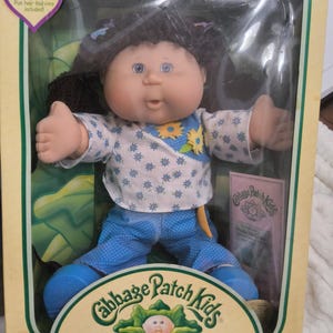 May include: A Cabbage Patch Kids doll with dark brown yarn hair, blue eyes, and a round face. The doll wears a white top with blue floral patterns and yellow flower accents, paired with blue polka-dot trousers and blue shoes, all inside a box.