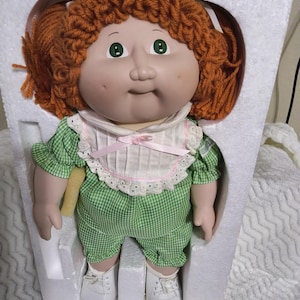 Könnte beinhalten: Eine Puppe mit orangefarbenem Wollhaar, grünen Augen und Grübchen in den Wangen. Die Puppe trägt einen grün-weiß karierten Strampler mit einem weißen Rüschenkragen mit rosa Schleifenverzierung sowie weiße Schnürschuhe in Styroporverpackung.