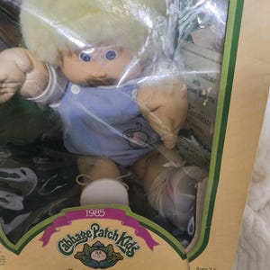 May include: A 1985 Cabbage Patch Kids doll with blonde yarn hair and blue eyes, wearing a light blue overall outfit with white buttons. The doll is packaged in a box featuring the text Cabbage Patch Kids and I'm unique. I'm adoptable.
