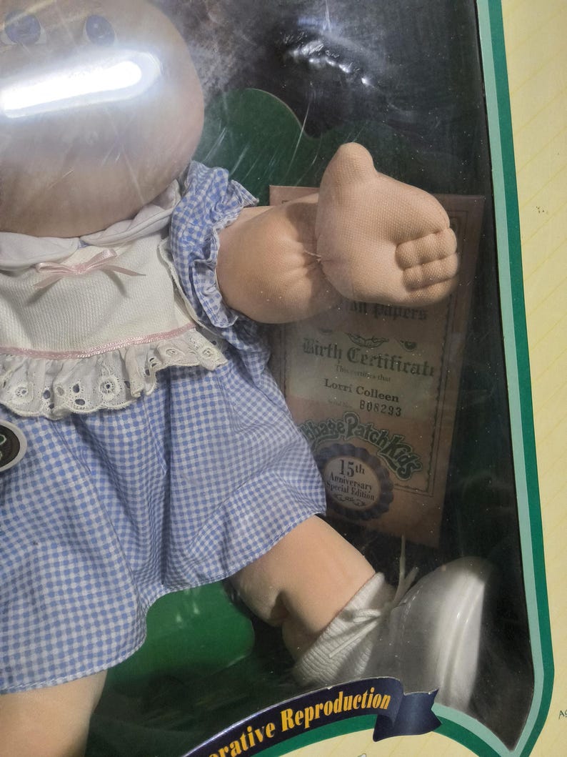 May include: A Cabbage Patch Kids doll with a round vinyl head and soft fabric body, wearing a blue and white gingham dress with a white lace-trimmed collar. The doll is in a box with a birth certificate labeled Lorri Colleen 808293 and 15th Anniversary.