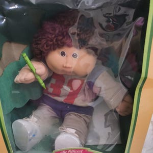 May include: A Cabbage Patch Kids doll with curly brown hair, wearing a grey and purple long-sleeved shirt with the number 39 printed in red, and grey pants. The doll holds a green crayon in its right hand and wears white shoes with blue accents.