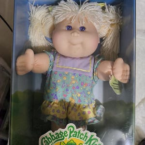 May include: A Cabbage Patch Kids doll with blonde yarn hair in pigtails, purple eyes, and a light skin tone. The doll wears a dress with a purple bodice and a patterned skirt featuring stick figures and hearts, packaged in a box.