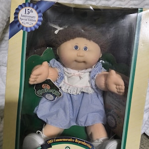 May include: A Cabbage Patch Kids 15th Anniversary Special Edition doll with brown yarn hair, blue eyes, and a light complexion. The doll wears a blue and white gingham dress with a white collar and white shoes, packaged in a cardboard box.
