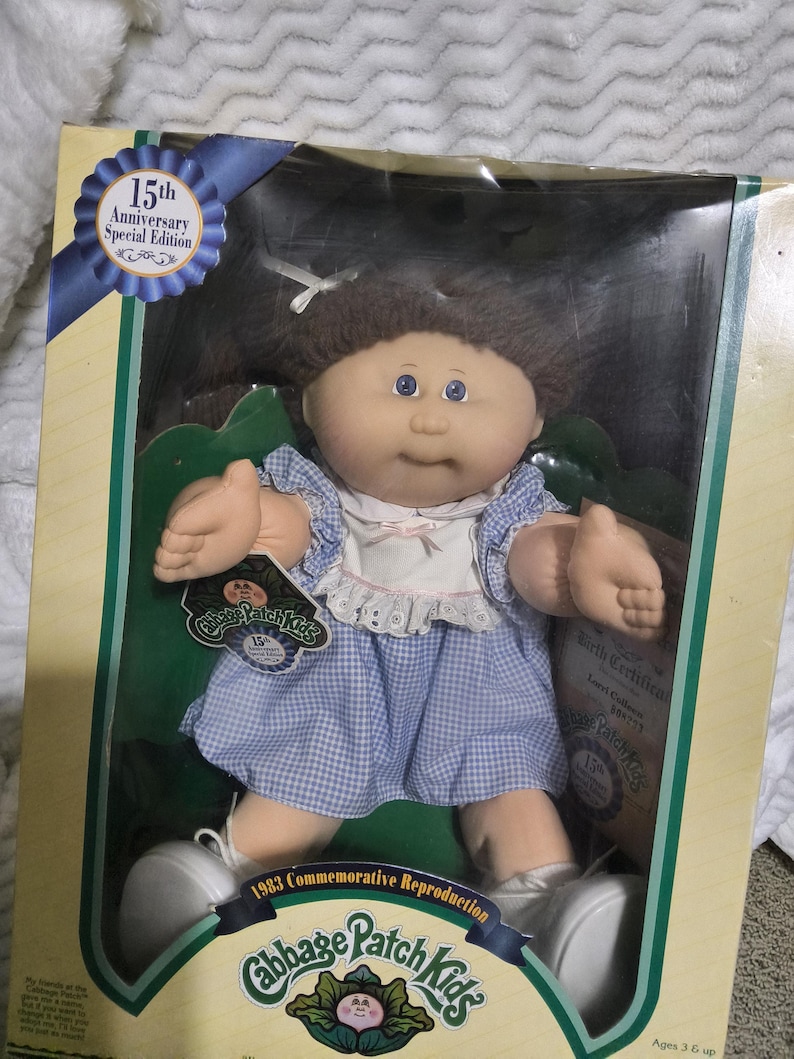 May include: A Cabbage Patch Kids 15th Anniversary Special Edition doll with brown yarn hair, blue eyes, and a light complexion. The doll wears a blue and white gingham dress with a white collar and white shoes, packaged in a cardboard box.