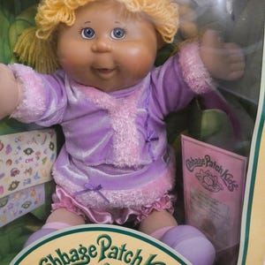 Könnte beinhalten: Eine Cabbage Patch Kids Puppe mit blondem Wollhaar und blauen Augen, bekleidet mit einer violetten Samtjacke mit rosa Kunstfellbesatz und passender Rüschenhose. Die Puppe befindet sich in der Originalverpackung, die einen Bogen Aufkleber enthält.