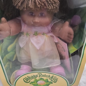 Op de afbeelding: Een Cabbage Patch Kids pop met bruin garen haar en paarse ogen, gekleed in een lichtroze jurk met een doorschijnende laag en roze schoenen. De pop zit in een kartonnen doos met het Cabbage Patch Kids logo en de tekst One of a Kind. And Just Mine!