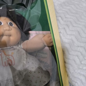 May include: A Cabbage Patch Kids doll with dark hair and a round face, wearing a white dress featuring a small orange giraffe graphic and a floral print skirt. The doll is packaged in a box with a visible certificate of authenticity.