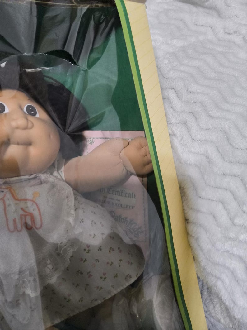 May include: A Cabbage Patch Kids doll with dark hair and a round face, wearing a white dress featuring a small orange giraffe graphic and a floral print skirt. The doll is packaged in a box with a visible certificate of authenticity.