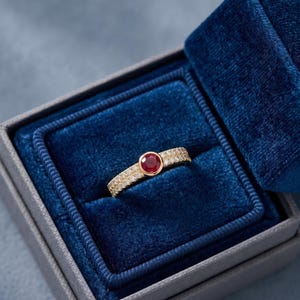 Può includere: Un anello in oro con un rubino rosso centrale a taglio rotondo in una montatura a castone, affiancato da due file di piccoli diamanti bianchi incastonati a pavé lungo la fascia, presentato in un portagioie foderato in velluto blu.