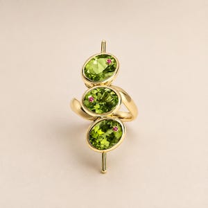 Könnte beinhalten: Ein Goldring mit drei vertikal angeordneten, oval geschliffenen Peridot-Edelsteinen, die jeweils mit einem kleinen, runden, leuchtend pinkfarbenen Edelstein akzentuiert sind. Das Goldband hat eine glatte, polierte Oberfläche und eine asymmetrische Form.