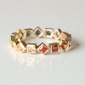 Könnte beinhalten: Ein goldener Ring mit einer durchgehenden Reihe quadratisch geschliffener Edelsteine in einem Farbverlauf aus Rot-, Orange-, Gelb- und Blautönen. Die Steine sind in einem sich wiederholenden Muster rund um den gesamten Metallring eingefasst.