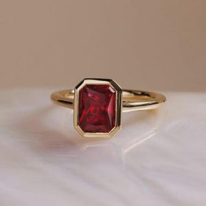 Könnte beinhalten: Ein Goldring mit einem rechteckigen, im Smaragdschliff geschliffenen, tiefroten Edelstein in einer glatten Zargenfassung. Die Ringschiene besteht aus poliertem Gelbgold mit abgerundetem Profil und zeigt ein klares, symmetrisches Design.