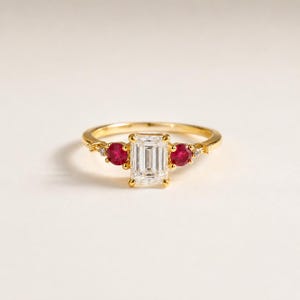 Könnte beinhalten: Ein Goldring mit einem zentralen, smaragdgeschliffenen, klaren Stein, der von zwei runden roten Steinen flankiert wird. Kleine klare Akzentsteine sind in die Ringschiene nahe der Mitte eingesetzt. Der Metallring ist dünn und poliert.