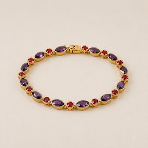 Op de afbeelding: Een goudkleurige armband met afwisselend marquise-geslepen paarse stenen en ronde rode stenen in een metalen ketting. Het sieraad is voorzien van een veilige baksluiting en vormt een cirkelvormig ontwerp met levendige kleuren.