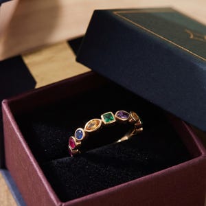 Könnte beinhalten: Ein Goldring mit einer Reihe verschiedenfarbiger Edelsteine in unterschiedlichen Schliffen, darunter rund, marquise und quadratisch. Der Ring befindet sich in einer dunklen Schmuckschatulle mit Klappdeckel und weichem, schwarzem Innenfutter.
