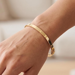 Puede incluir: Una pulsera rígida articulada de tono dorado pulido con una superficie plana y lisa y un mecanismo de cierre seguro. La pulsera presenta un acabado metálico reflectante que capta la luz, mostrando un diseño de banda limpia y continua.