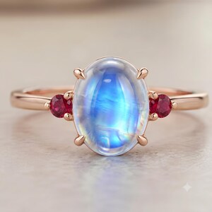 Könnte beinhalten: Ein Ring aus Roségold mit einem großen, ovalen, irisierenden Mondstein-Cabochon in der Mitte, flankiert von zwei kleinen, runden, leuchtend roten Rubinen. Die Steine werden von feinen Metallkrappen auf einer glatten, polierten Ringschiene gehalten.