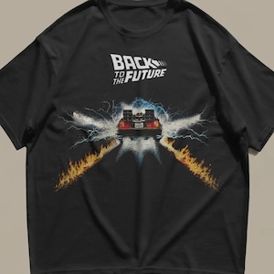 May include: A black short-sleeved t-shirt featuring the Back to the Future logo in white text at the top. Below the text, a graphic depicts a car driving away, surrounded by blue electrical energy bolts and two trails of orange fire on the ground.