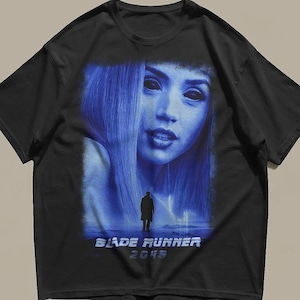 以下が含まれることがあります： 黒の半袖Tシャツ。青を基調とした大きなグラフィックプリントが施されており、暗い目の人物の顔と、その下に立つ小さな人物のシルエットが描かれています。グラフィックの下部には「BLADE RUNNER 2049」という文字が白でプリントされています。