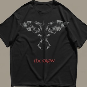 以下が含まれることがあります： 黒の半袖Tシャツで、グレーと白の色合いで翼を広げた様式化された鳥のグラフィックが描かれています。グラフィックの下には、胸の中央に太字の赤いセリフ体で「THE CROW」というテキストがプリントされています。