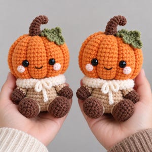Puede incluir: Dos figuras de calabaza de ganchillo idénticas con cuerpos naranjas, tallos marrones y hojas verdes. Cada figura tiene ojos de cuentas negras, mejillas rosadas, una pequeña sonrisa, un cuello color crema con lazo y manos y pies marrones.
